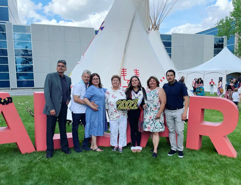 Seven people stand outside a tipi, the woman in the middle wears graduation regalia and holds a sign reading '2022.'